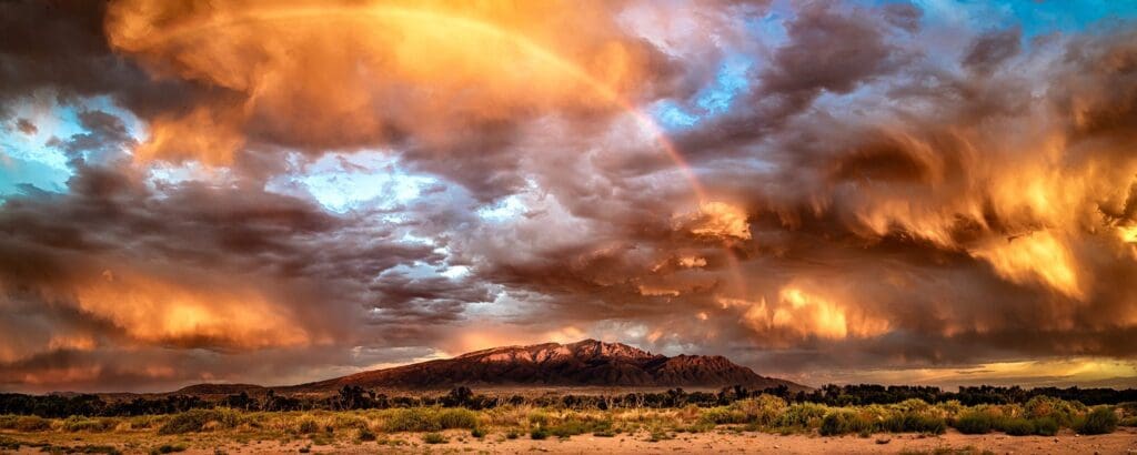 A dramatic sky with a vibrant rainbow arcs over a mountain landscape. The clouds are illuminated with fiery shades of orange and gold from the setting sun, casting a warm glow over the scene. Desert terrain with sparse vegetation is in the foreground.