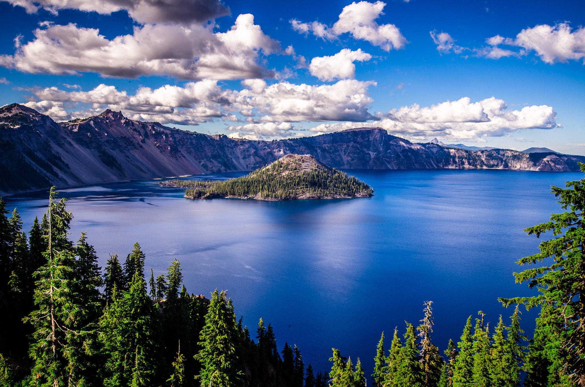 A bright, clear day at Crater Lake, with deep blue water surrounded by rocky cliffs. Lush greenery in the foreground and an island in the center. White, fluffy clouds scatter across the vibrant blue sky.