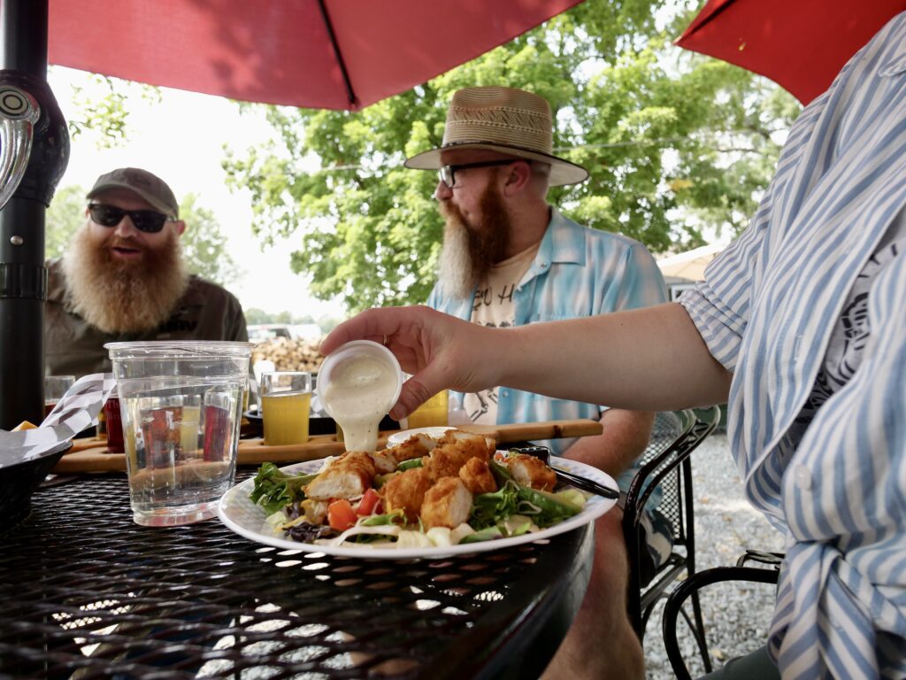 Three people sit at an outdoor table under red umbrellas. In the foreground, a hand pours dressing onto a salad plate. Two men with beards, sunglasses, and hats sit in the background, talking and smiling.