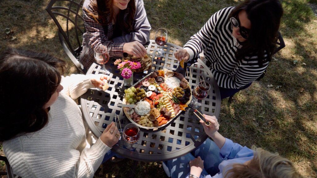 Four women sit around a lattice outdoor table sharing a charcuterie board with fruit, cheeses, and crackers. Each has a glass of red wine, and they are enjoying a sunny meal together outside.