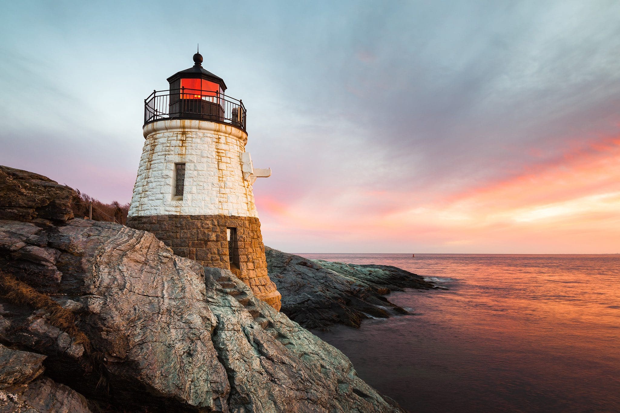 A lighthouse stands on a rocky shoreline at sunset, with a dramatic sky of pink, orange, and purple hues. The sea is calm, reflecting the colorful sky. The lighthouse is made of white and stone brick.