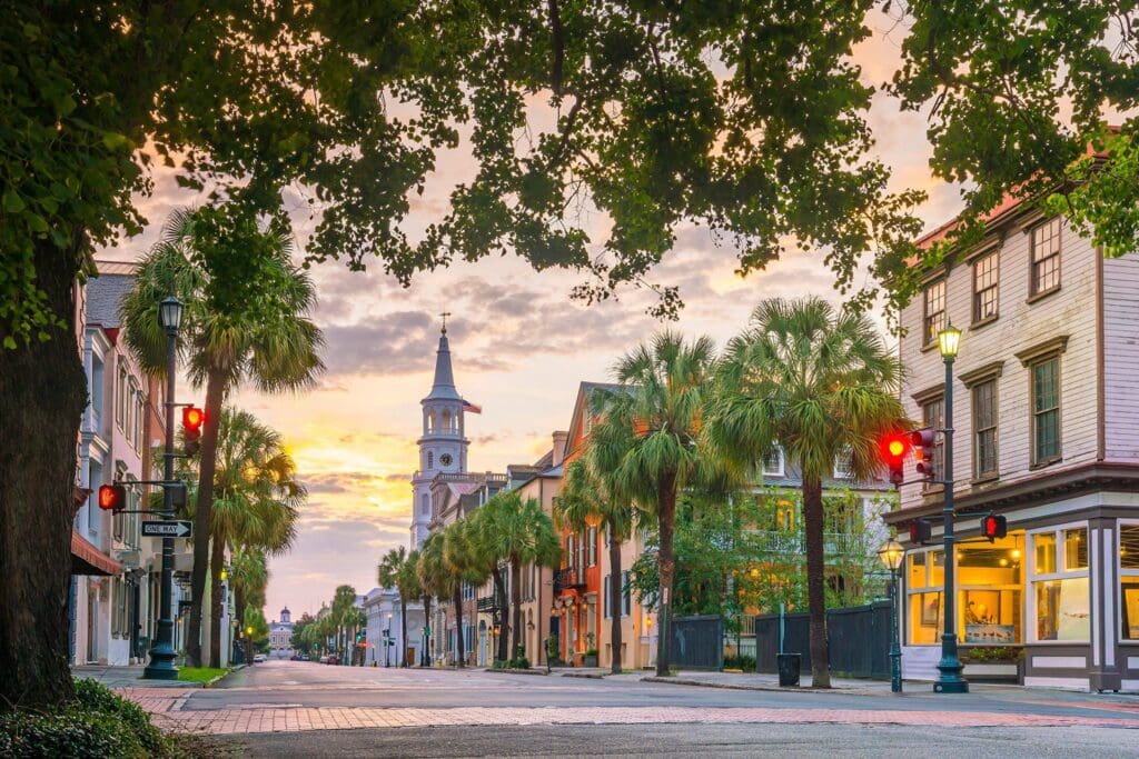 A picturesque street scene with historic buildings, palm trees lining the road, and a prominent church steeple in the background. The sky is partly cloudy with warm sunset hues. Streetlights and a quiet intersection complete the charming atmosphere.