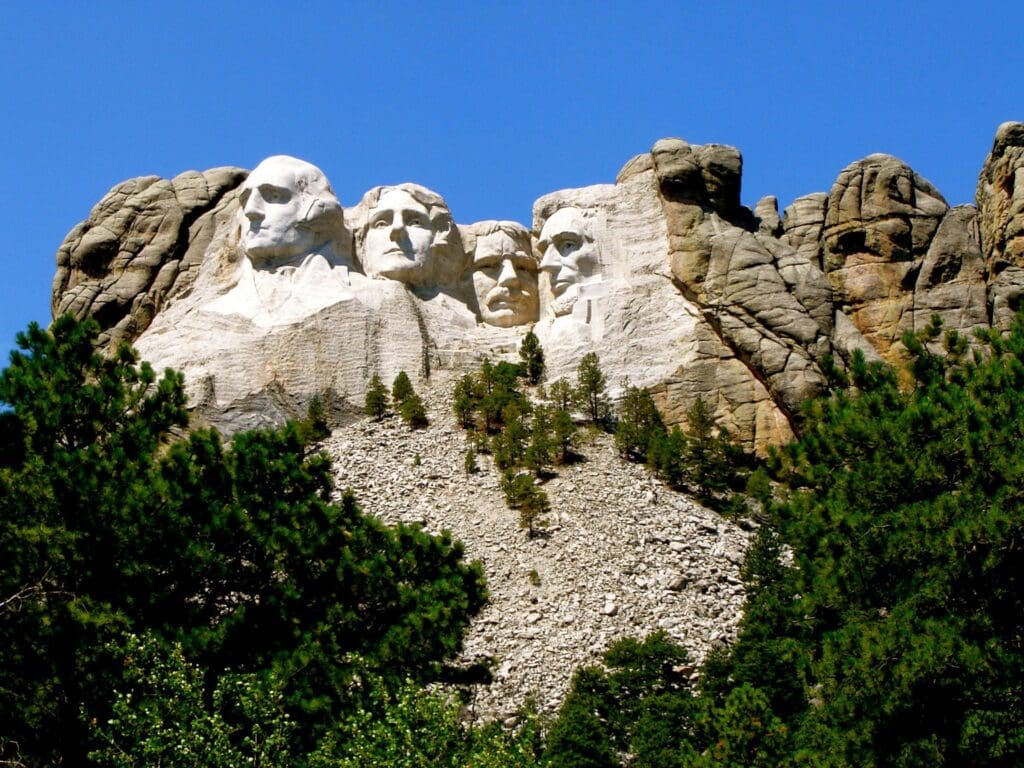 Mount Rushmore National Memorial featuring the carved faces of four U.S. presidents: George Washington, Thomas Jefferson, Theodore Roosevelt, and Abraham Lincoln. Surrounded by trees, under a clear blue sky.
