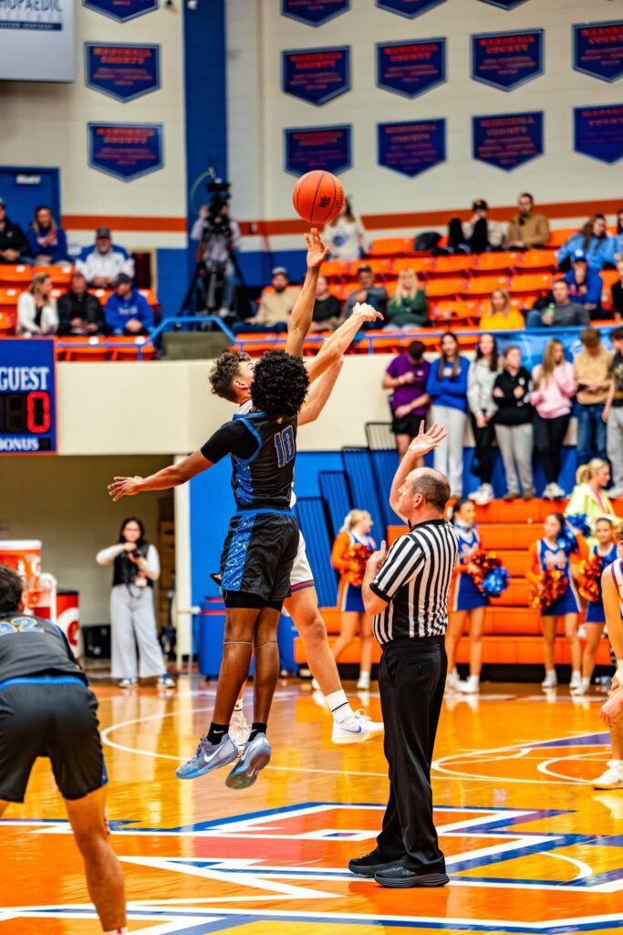Two basketball players jump for the ball at tip-off, as a referee watches. One player wears black and blue, number 10. Spectators and cheerleaders are visible in the background on orange bleachers.