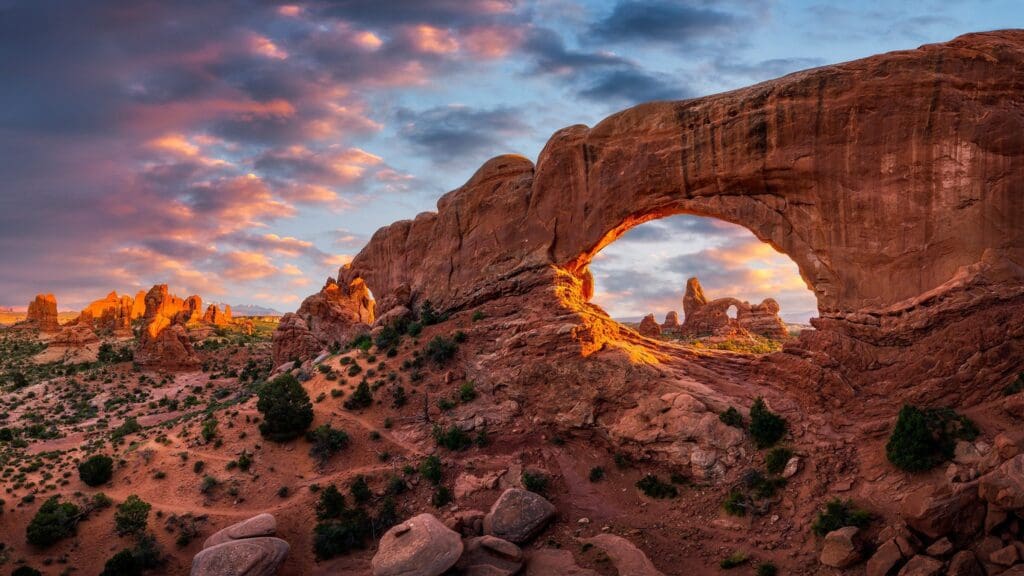 Sunset view of a large sandstone arch in a desert landscape. The arch frames a smaller rock formation in the distance. The sky is vibrant, with orange clouds, and the landscape features scattered shrubs and rocks.