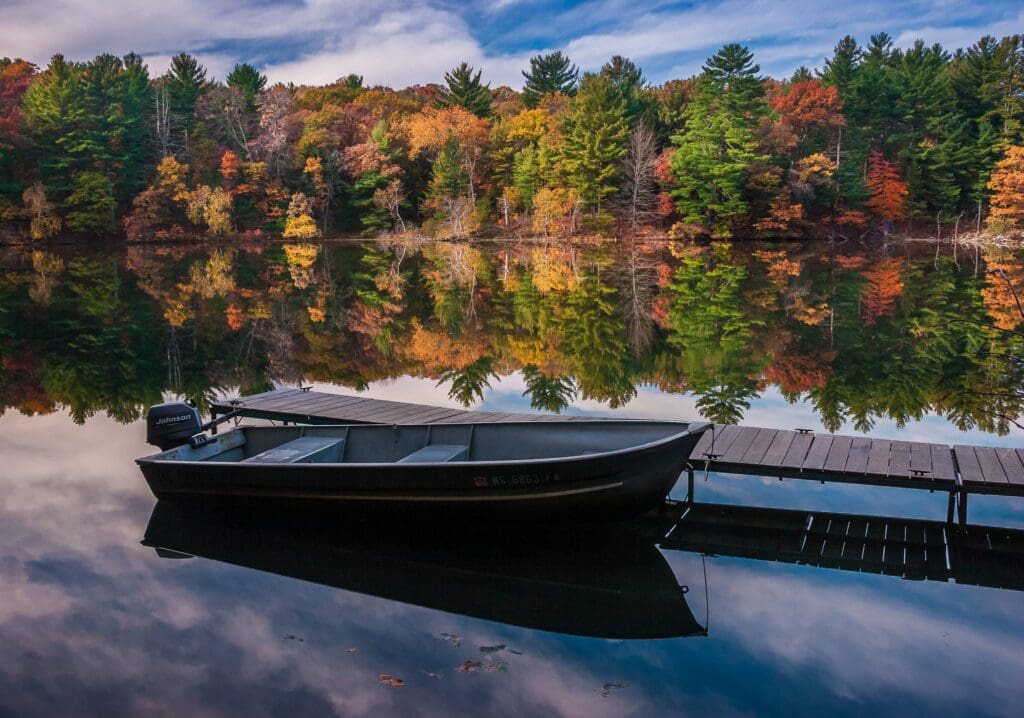 A small motorboat is tied to a wooden dock on a calm lake. The water reflects vibrant autumn trees with red, orange, and green leaves under a partly cloudy sky. The scene captures the serene beauty of fall.