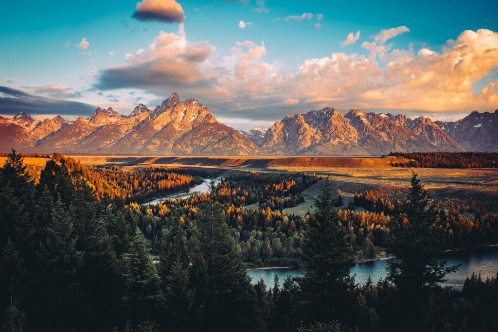 A scenic view of a mountain range during sunset. The sky is partly cloudy with warm, golden hues illuminating the peaks. A winding river flows through a lush forest in the foreground, with dark green pine trees in the foreground.