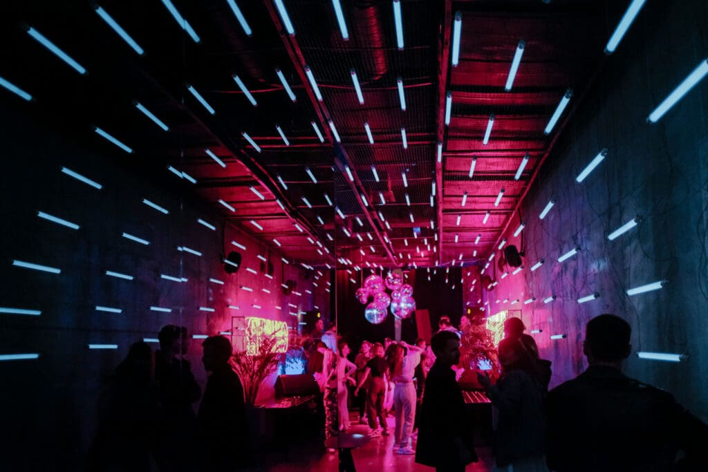 A dimly lit room features neon blue and pink lights on the walls and ceiling. Several people stand and converse near the entrance, where disco balls hang, creating a vibrant, party atmosphere.