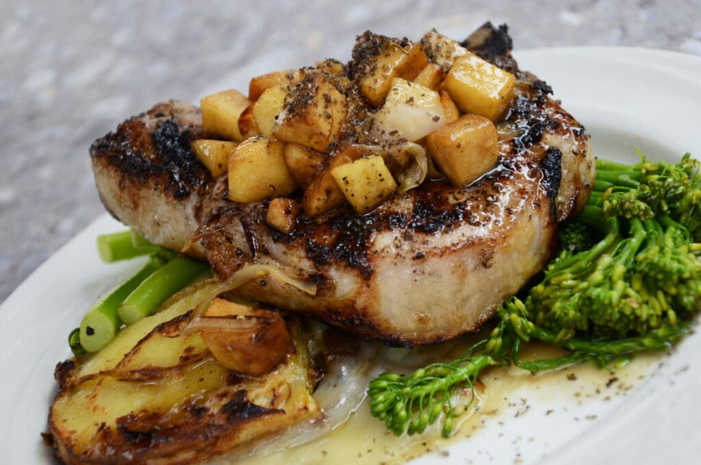 A grilled pork chop topped with diced apples and onions is served on a white plate. The dish is accompanied by broccolini and roasted potatoes, all drizzled with a savory sauce.