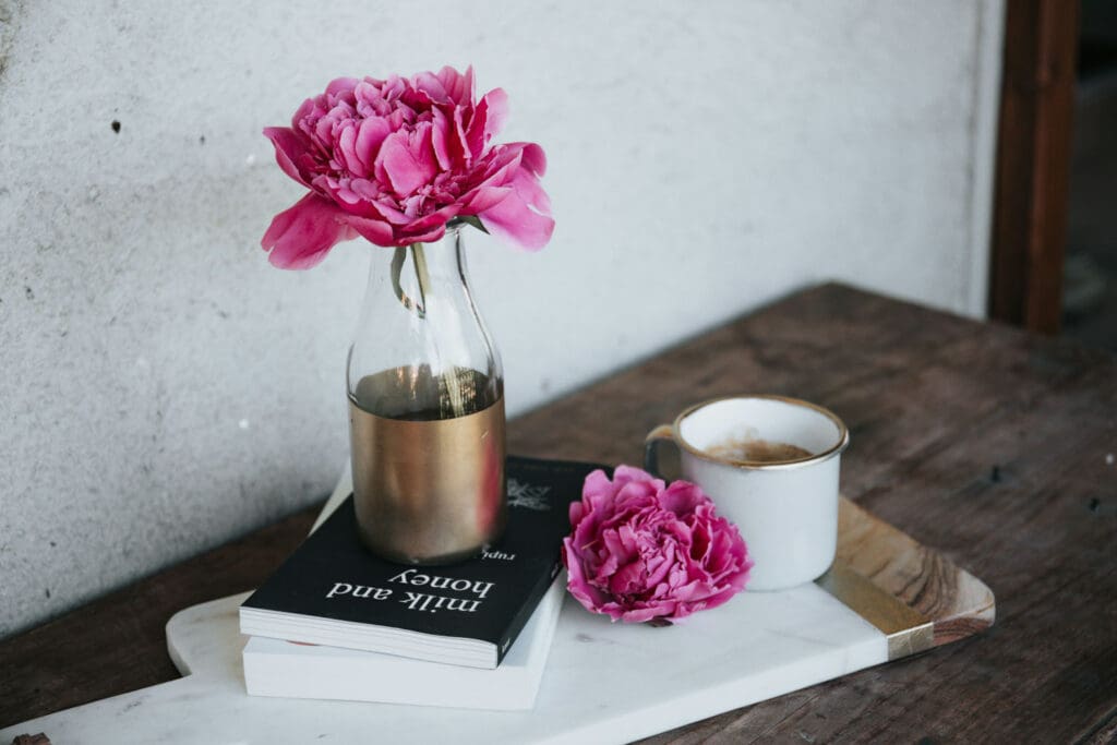 A pink peony in a glass vase sits on a book titled Milk and Honey on a wooden table. Beside it is a white cup filled with coffee and a loose pink peony blossom, all arranged on a white tray.