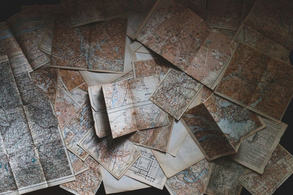 A scattered collection of vintage maps, featuring various geographic regions. The maps display intricate details, including rivers, roads, and topographic lines, in muted earth tones. Some maps are folded, while others lie flat on a dark surface.