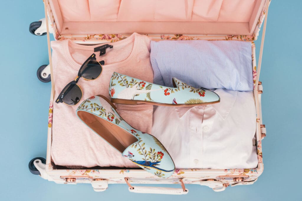 An open suitcase on a light blue background contains folded clothes, including a pink shirt, a blue striped shirt, and a white shirt. A pair of floral-patterned shoes and sunglasses are placed on top.