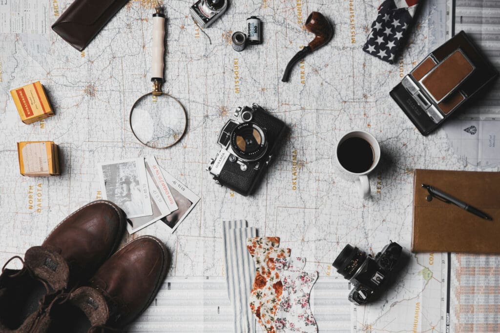 A flat lay of vintage travel items on a map: a camera, magnifying glass, pipe, coffee cup, notebook with pen, colorful photos, boots, film boxes, a vintage camera, and a floral handkerchief. American flag pattern visible on fabric in the corner.