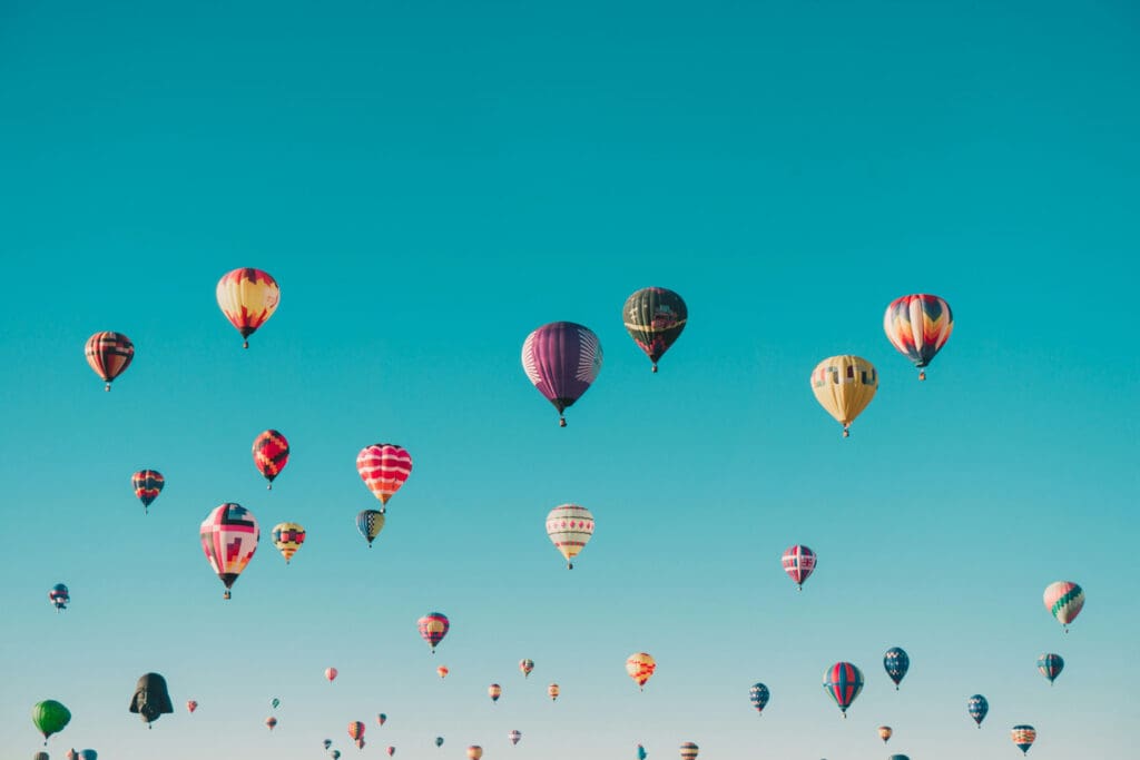 A clear blue sky filled with numerous colorful hot air balloons, each featuring different patterns and designs. The balloons are spread across the horizon, creating a vibrant and cheerful scene.