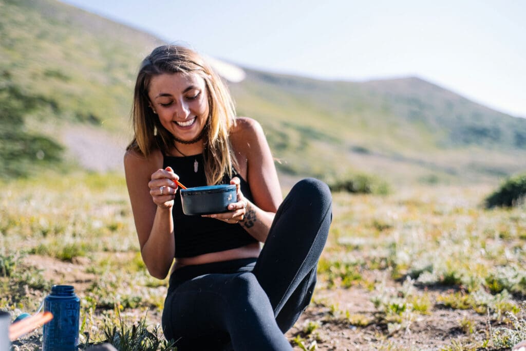 A woman sitting on grassy terrain smiles while eating from a container with a red spoon. She is in a mountainous landscape under a clear sky, wearing a black tank top and leggings. A blue water bottle is nearby.