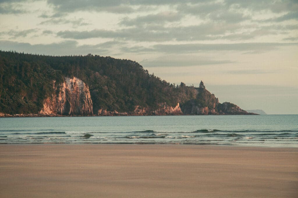 A serene beach scene with gentle waves lapping at the sandy shore. In the background, a lush, tree-covered hill with rugged cliffs under a cloudy sky. The light has a warm, soft glow, suggesting early morning or late afternoon.