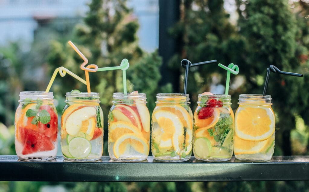 Five mason jars filled with refreshing fruit-infused water sit on a ledge. Each jar contains colorful fruit slices such as watermelon, lime, orange, and berries, with straws in various colors against a leafy, blurred background.