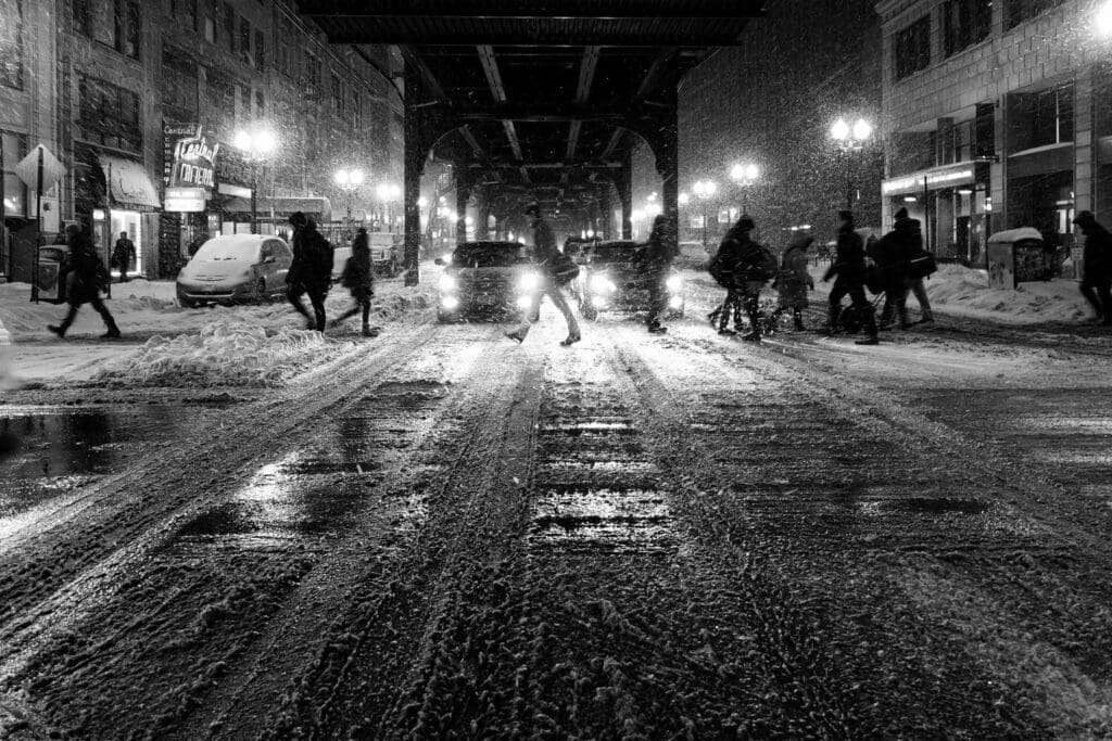 Black and white photo of a snow-covered city street at night. Cars have headlights on, casting light on pedestrians crossing. Buildings with lit signs line the street, and snow is falling, creating a reflective, wintry atmosphere.