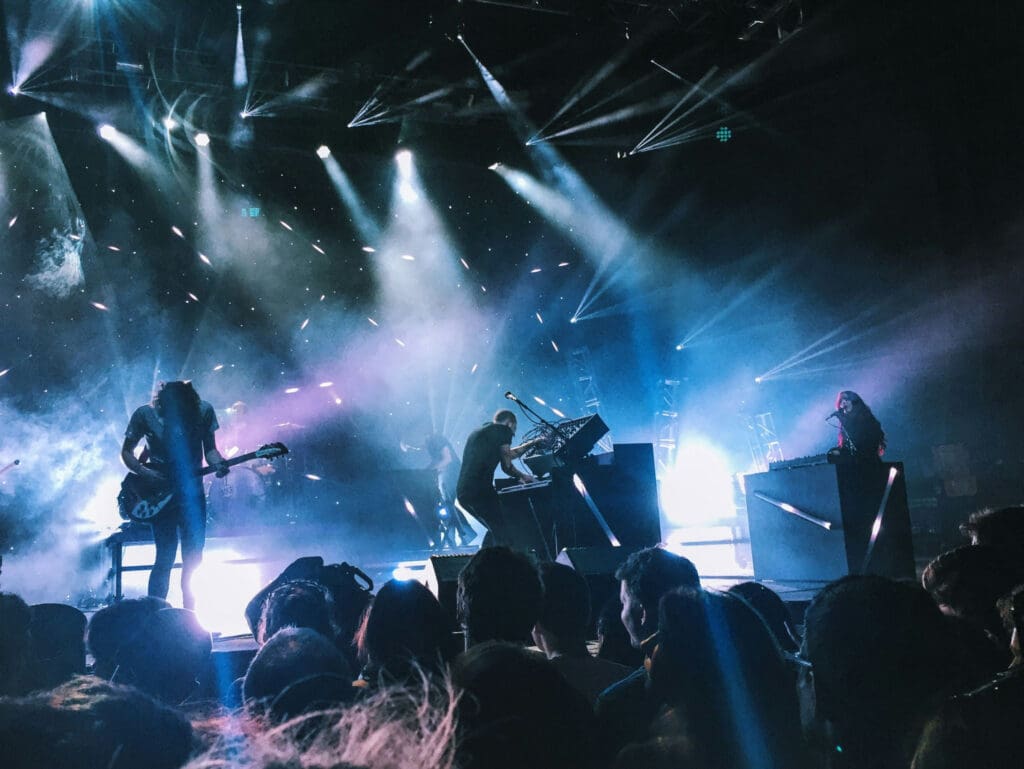 A band performs on a dimly lit stage with colorful lights and smoke effects. Silhouettes of the musicians and their instruments are visible against the vibrant backdrop, while an audience watches from the foreground.
