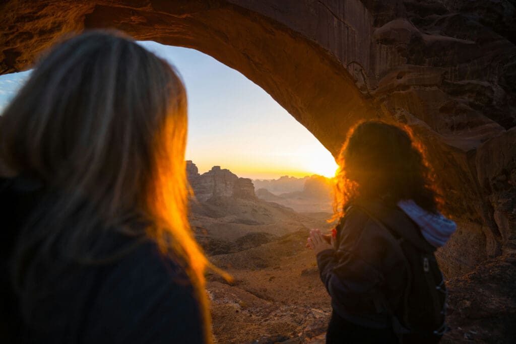 Two people stand under a large rock arch, facing a scenic view of a mountainous desert landscape at sunset. The sun casts a warm glow, silhouetting the figures and the surrounding terrain.