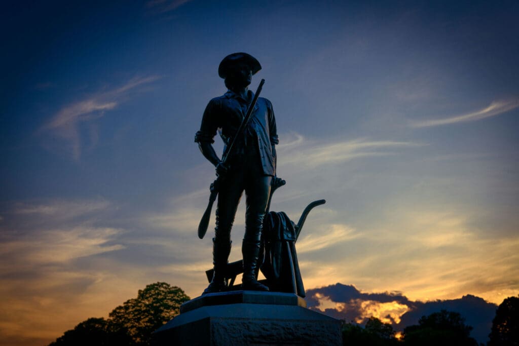 Silhouette of a statue featuring a soldier wearing a hat and holding a rifle, standing on a pedestal. The background displays a dramatic sunset with orange, yellow, and blue hues in the sky.