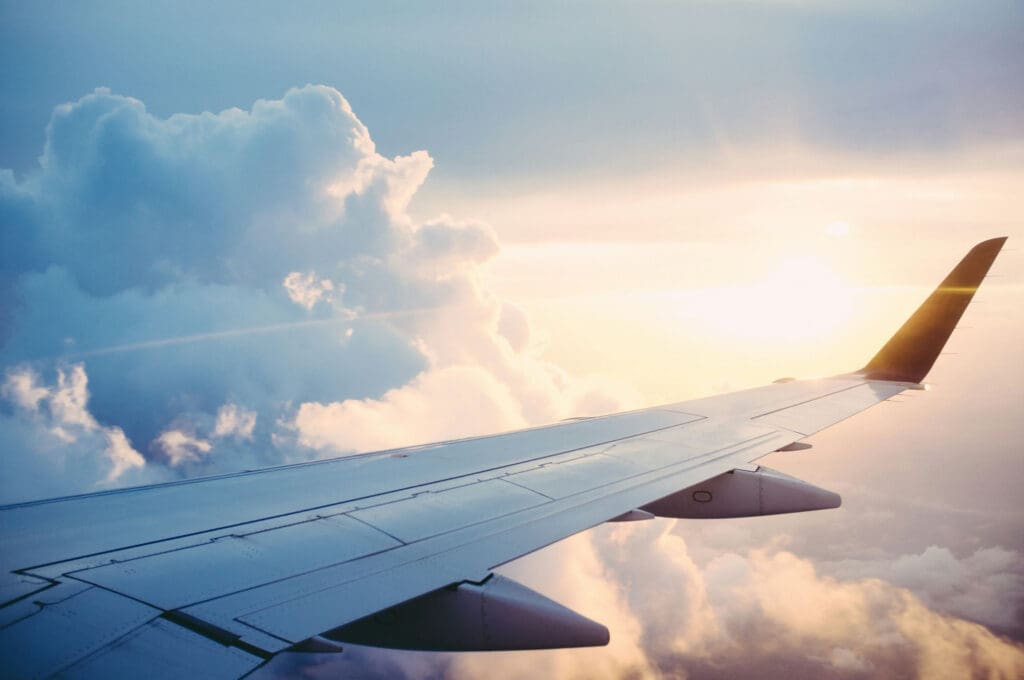 View from an airplane window showing the wing and a sunny sky with fluffy clouds. The sun casts a warm glow, illuminating the clouds and sky in soft shades of orange and blue.