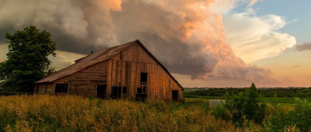 A rustic wooden barn stands amidst tall grasses under a dramatic sky. Thick, colorful clouds swirl above, casting an ethereal glow over the rural landscape, with hints of a sunset in the background.