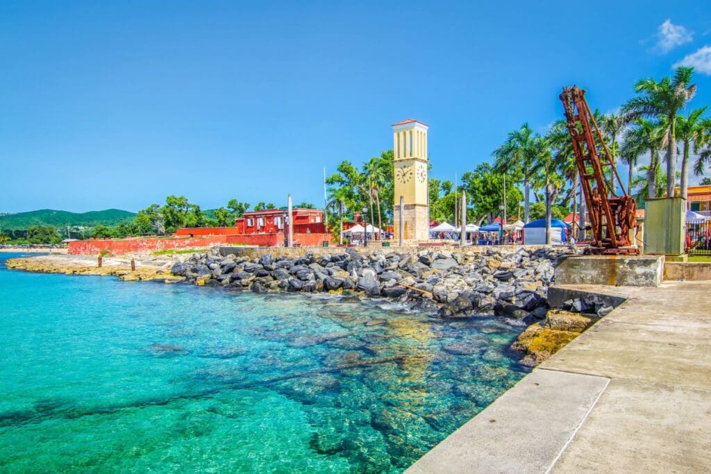 A vibrant coastal scene with clear turquoise water, a rocky shoreline, and a clock tower flanked by palm trees. Red buildings and tents are visible in the background under a bright blue sky.