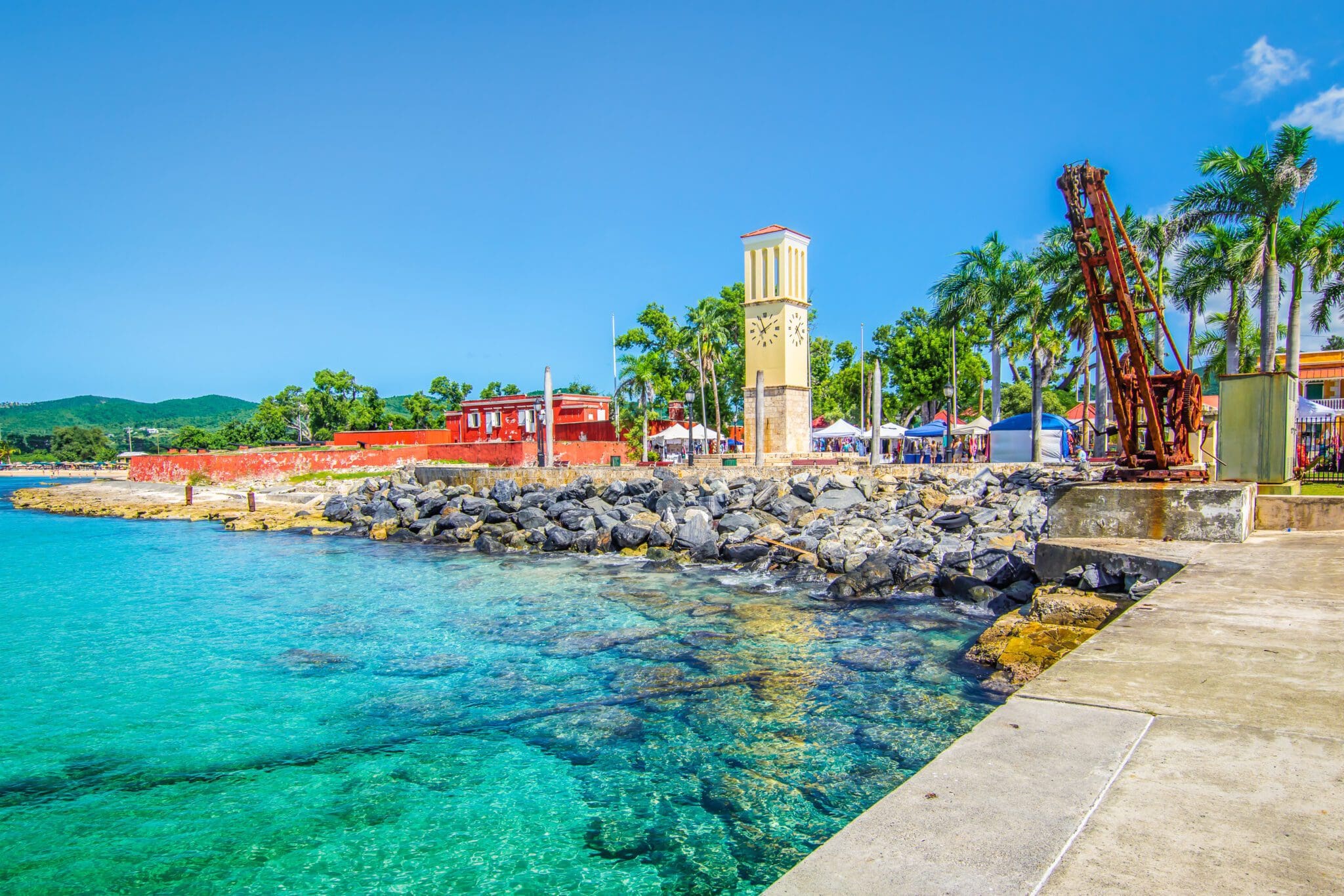 A vibrant coastal scene with clear turquoise water, a rocky shoreline, and a clock tower flanked by palm trees. Red buildings and tents are visible in the background under a bright blue sky.