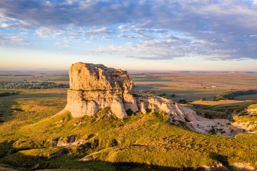 A large rock formation stands prominently in a grassy landscape under a partly cloudy sky. The structure is illuminated by warm, golden sunlight, casting soft shadows. The horizon stretches into fields and distant flatlands.