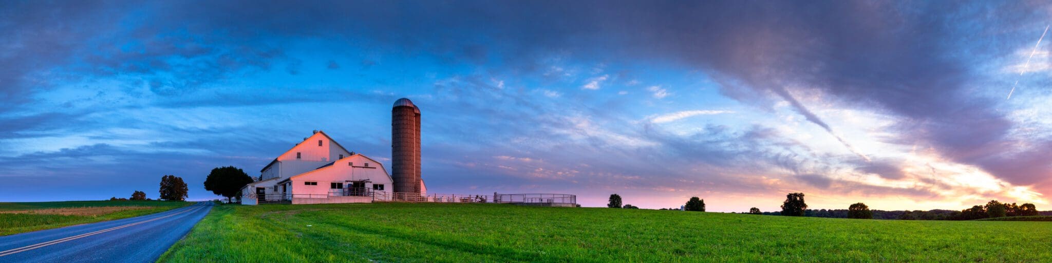 A panoramic view of a rural landscape at sunset, featuring a white barn with a tall silo on the left. The sky is a mix of blue, pink, and orange hues, with wispy clouds. A road runs alongside a green, open field.