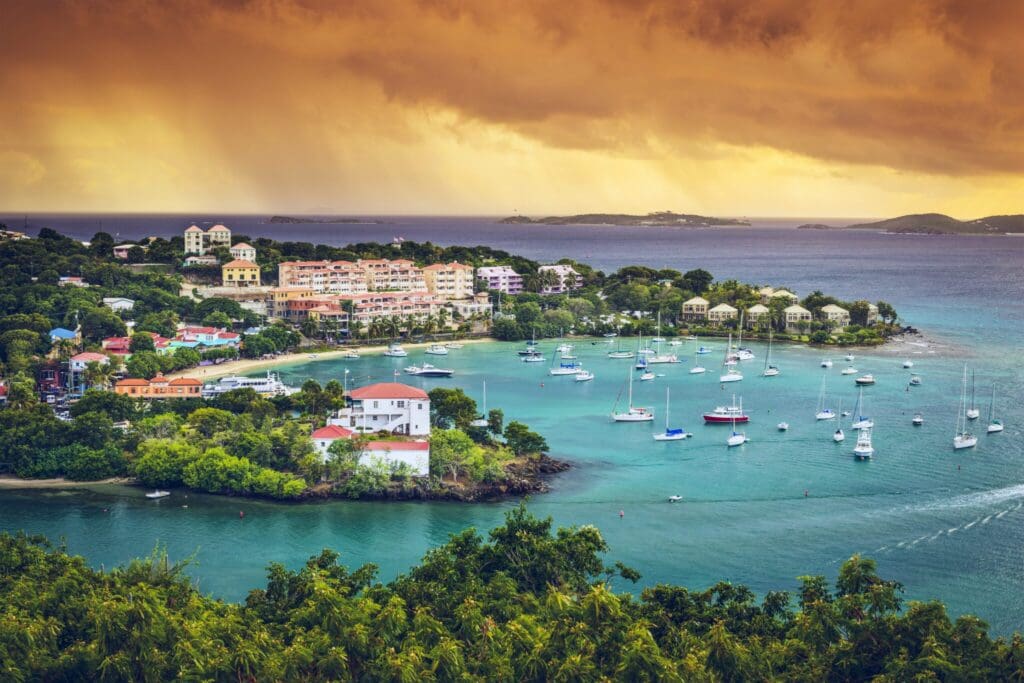 A scenic coastal view of a small harbor, surrounded by lush greenery and colorful buildings. Numerous sailboats float on calm turquoise waters beneath an orange-tinted cloudy sky, suggesting a sunset or sunrise.