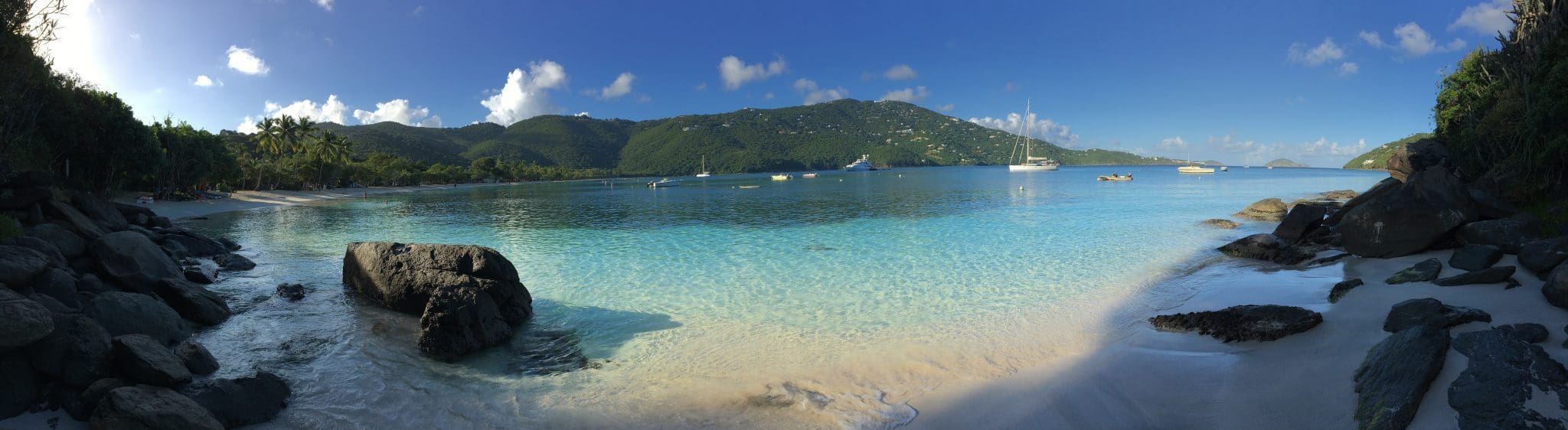A serene beach scene with clear turquoise water, rocky shores, and distant green hills under a blue sky. Boats float on the water, and a few clouds scatter across the sky. The sun casts a gentle light on the landscape.
