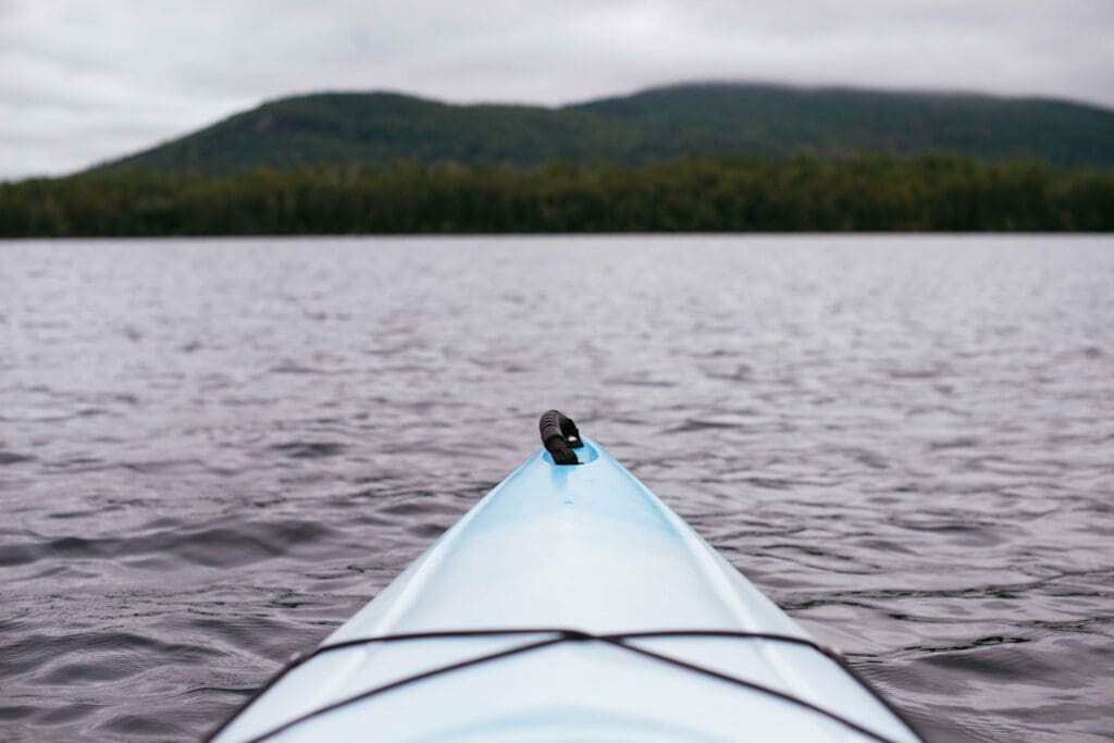 Front view of a blue kayak gliding on a calm lake, with forested hills and a cloudy sky in the background.