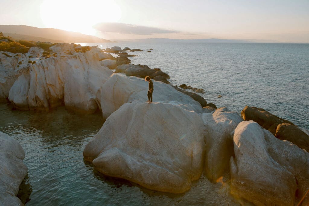 A person stands on large, smooth rocks overlooking a calm sea at sunset. The sky is clear with a few clouds, and the sun casts a warm glow over the scene. Rocky shorelines and distant mountains are visible in the background.