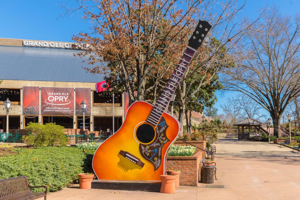 A large decorative guitar stands outside the Grand Ole Opry venue. The guitar is orange and decorated with floral designs. Nearby, there are trees, and banners with the Grand Ole Opry name hang on the building. Its a clear day.