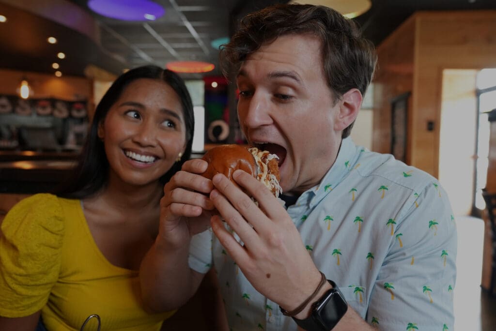 A man eagerly bites into a large sandwich while sitting in a restaurant. A woman next to him smiles and watches. The setting has modern decor with colorful overhead lights. The man wears a palm tree patterned shirt, and the woman wears a yellow top.
