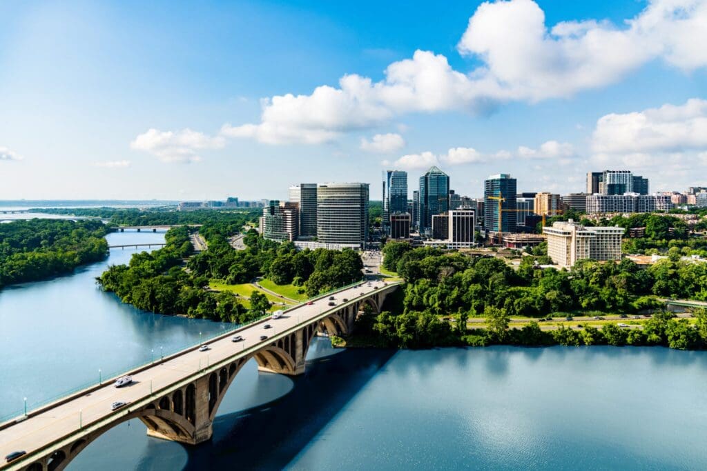 Aerial view of a cityscape with a bridge spanning a river. Tall, modern buildings rise against a bright blue sky with scattered clouds. Lush greenery surrounds the water, and vehicles are seen crossing the bridge.