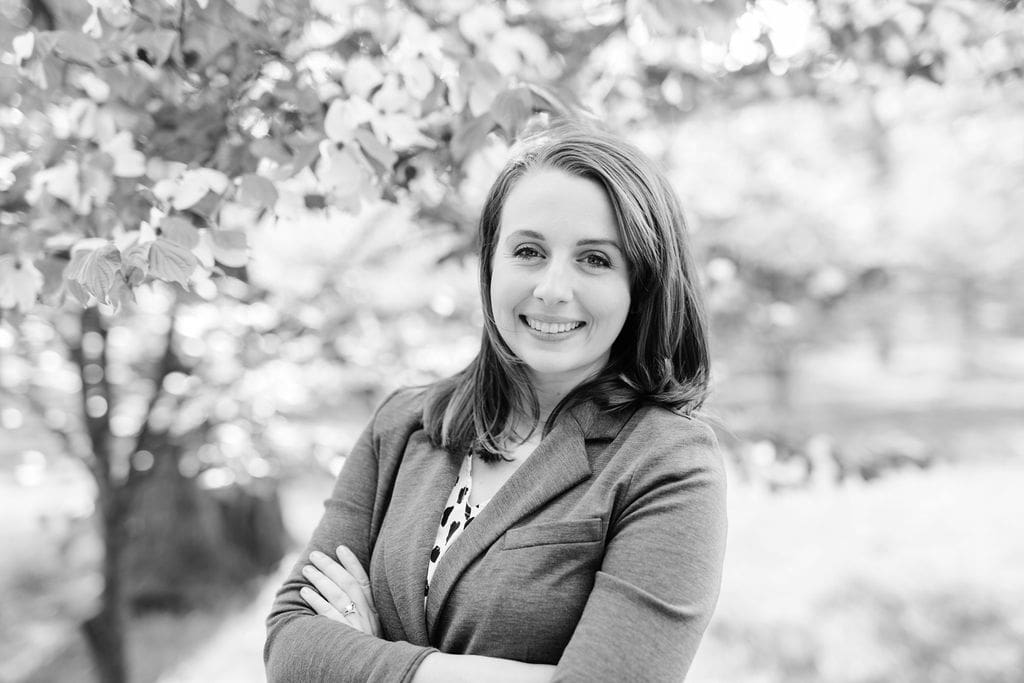 A woman with long hair is smiling and standing with her arms crossed. Shes wearing a blazer and patterned top. The background is an outdoor setting with trees in soft focus. The image is in black and white.