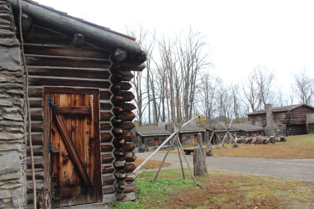 A historic site featuring several log cabins with weathered wood exteriors. The foreground shows a cabin with a wooden door. Bare trees stand in the background under a cloudy sky. The ground is a mix of grass and gravel.