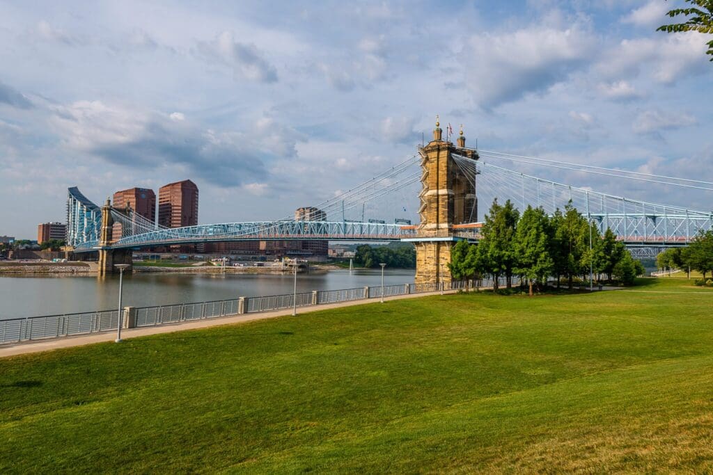 A large suspension bridge spans a calm river, connecting two urban areas. The bridge features two stone towers and blue cables. The foreground includes a grassy park with trees, while red-brick buildings are visible in the background under a partly cloudy sky.