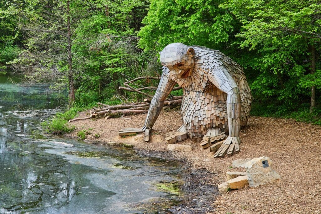 A large wooden sculpture of a troll, sitting by a pond in a forest setting. The troll has a hunched posture, with one hand touching the water. Green trees surround the scene, and there are small branches nearby.
