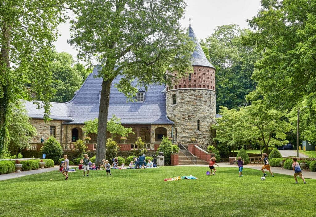 A group of children play soccer on a lush green lawn in front of a charming stone building with a turret and arched windows. The scene is set amidst tall trees and vibrant greenery under a clear sky.
