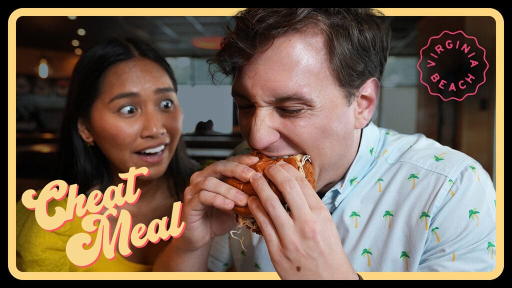 A man eagerly bites into a large sandwich while a woman next to him looks surprised and amused. The words Cheat Meal and Virginia Beach are displayed in stylized text. They are sitting in a restaurant setting.