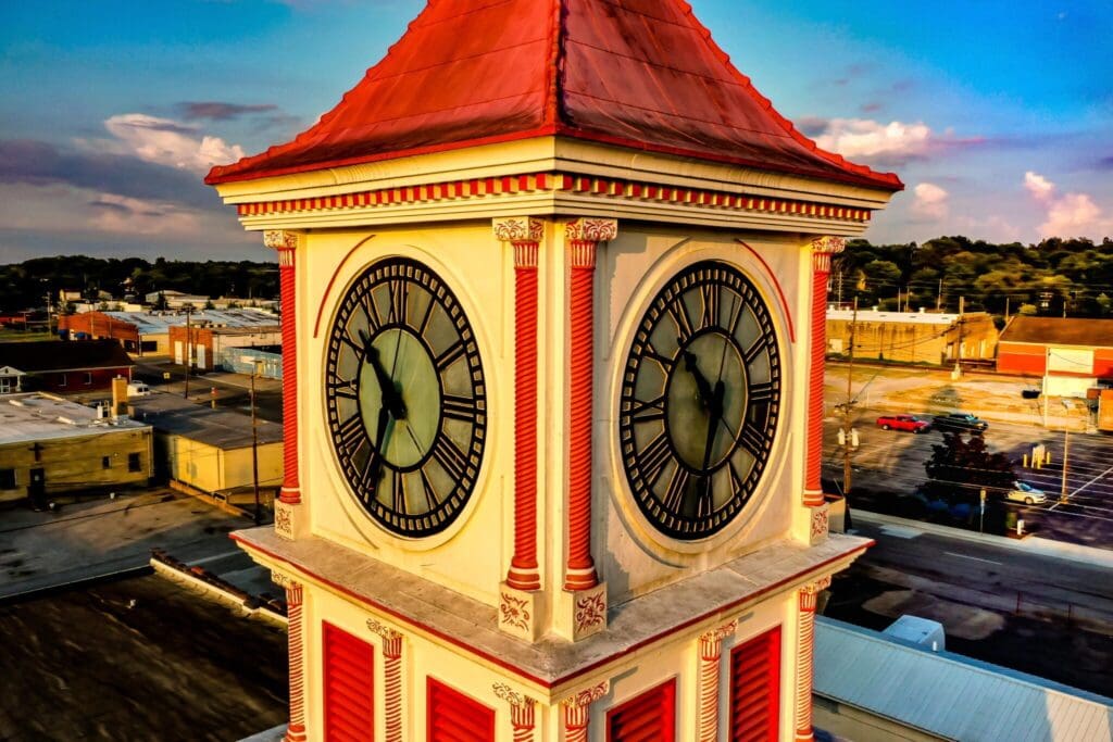 Aerial view of a clock tower with a red roof and decor against a backdrop of a small town. The clock face shows the time at approximately 6:00. The sky is partly cloudy with a warm, golden hue.