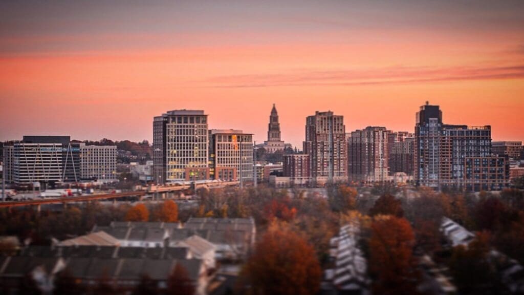 Cityscape at sunset with a skyline of various modern high-rise buildings and a prominent tower in the background. The foreground features a residential area with houses, and the scene is enveloped in warm, soft orange and pink hues.