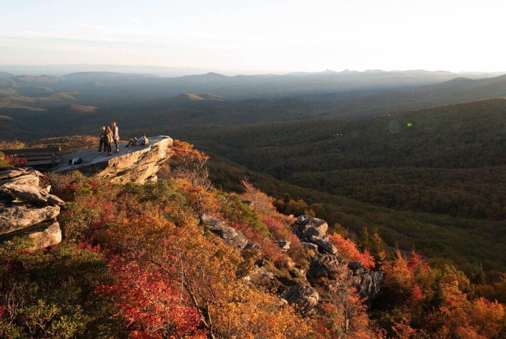 Four people stand on a rocky ledge overlooking a vast landscape of rolling hills covered in autumn foliage, with hues of orange and red. The sun is low, casting warm light across the scene.
