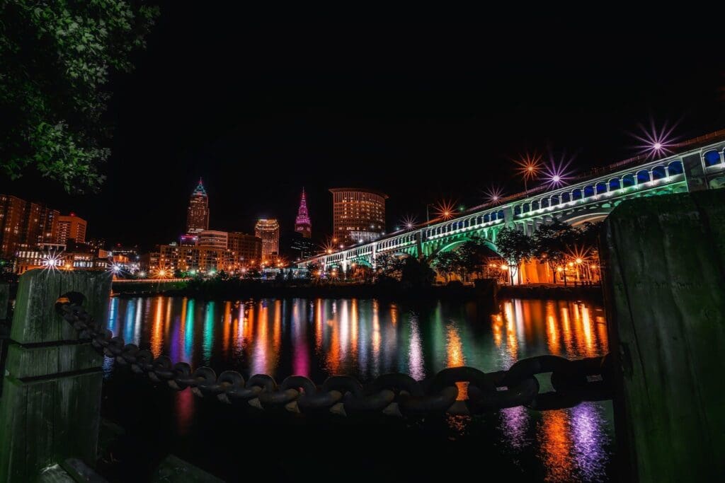 Night view of a cityscape with a brightly lit bridge and skyscrapers reflecting in a calm river. Colorful lights create a vibrant atmosphere. A large chain and wooden post are visible in the foreground, adding depth to the scene.