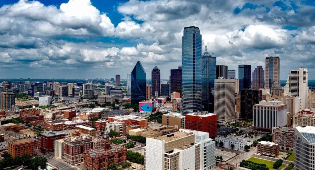Aerial view of a city skyline with tall skyscrapers under a partly cloudy sky. The foreground features a mix of modern and historic buildings, and the scene includes greenery scattered throughout the urban landscape.