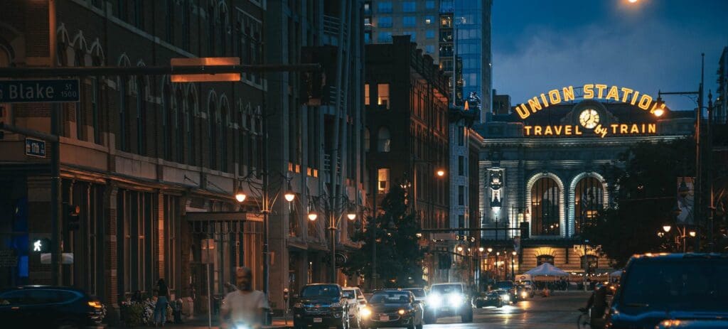A nighttime street view leads toward the illuminated Union Station building. Neon signs read Union Station Travel by Train. The street is bustling with cars and bicycles, and the sky is a deep blue, contrasting with the warm streetlights.